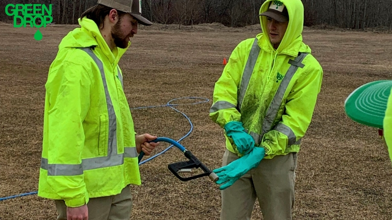 Green Drop employees in high-visibility jackets preparing a deep-root fertilization tool for lawn treatment on a dry field.