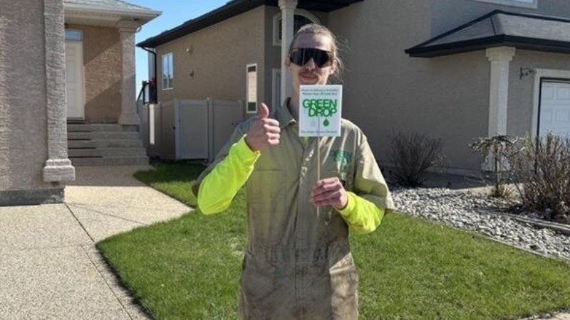 A Green Drop professional giving a thumbs-up while holding a Green Drop sign in front of a freshly maintained lawn in a residential area.