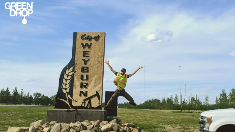 Green Drop worker jumping in front of a Weyburn city sign